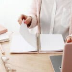 A person wearing a white blouse and pink cardigan sits at a desk, flipping through pages of a 30 day guided journal. Nearby are a pink pen, eyeglasses, closed notebooks, and a laptop.