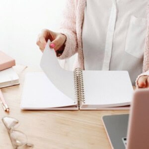 A person wearing a white blouse and pink cardigan sits at a desk, flipping through pages of a 30 day guided journal. Nearby are a pink pen, eyeglasses, closed notebooks, and a laptop.