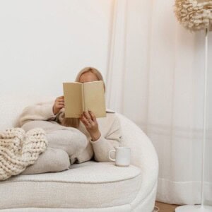 A person sits on a cream-colored sofa, wrapped in a knit blanket, reading about journaling myths. A white mug rests beside them. The setting is cozy with soft, neutral tones and a floor lamp in the background.