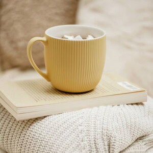 A yellow textured mug filled with marshmallows sits atop an open book—a perfect scene for any good writer—resting on a folded, cream-colored knitted blanket. The softly blurred background creates a cozy atmosphere.