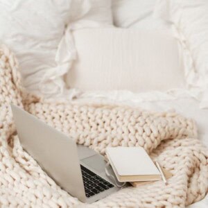 A silver laptop, journaling supplies like an open notebook and pencil rest on a chunky cream knit blanket with white pillows in the background, creating a cozy workspace setting.