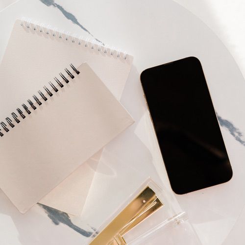 Essential Journaling Supplies for Beginners 2 Two spiral notebooks, a black smartphone, and a gold pen in a clear case—perfect journaling supplies—are arranged on a white, marble-patterned surface in bright, natural light.