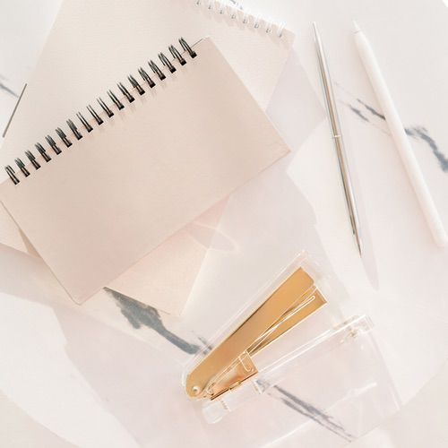 Essential Journaling Supplies for Beginners 3 An overhead view of a workspace with journaling supplies, including two spiral notebooks, a silver pen, a white pencil, and a clear stapler with gold components arranged on a white marble surface.