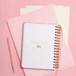 A spiral notebook with the words dream. Plan. Do. On the cover, perfect for guided journaling, rests atop pink folders beside a pink pencil, all arranged on a pink background.