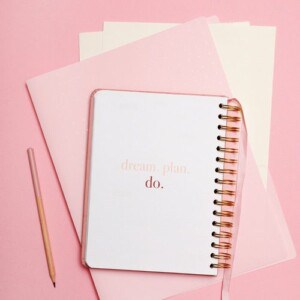 A spiral notebook with the words dream. Plan. Do. On the cover, perfect for guided journaling, rests atop pink folders beside a pink pencil, all arranged on a pink background.