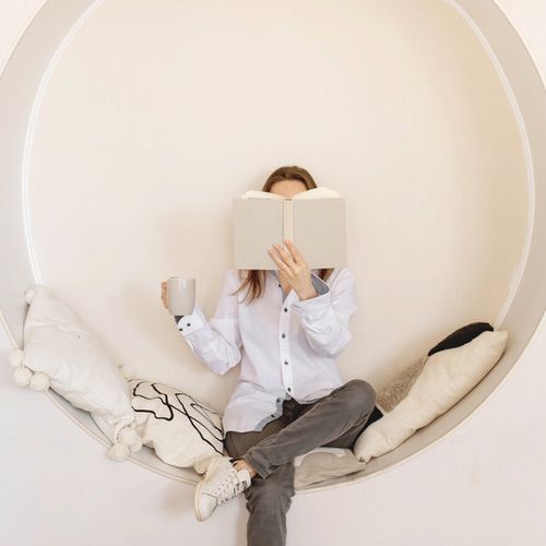 A person sits in a round, built-in wall nook with pillows, holding a mug in one hand and reading an open book that covers their face—perhaps enjoying insights from a headway app review. They wear a white shirt and gray pants.