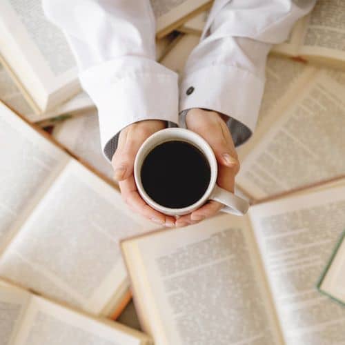 Headway App Review 5 A person in a white shirt holds a mug of black coffee above a table covered with open books, perhaps pausing to reflect on reading a headway app review, viewed from above.