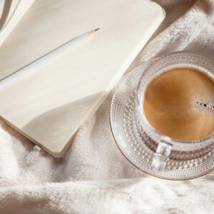 A clear glass cup of coffee sits on a matching saucer next to an open blank notebook—your next best journal—and a white pencil, all resting on a soft, cream-colored fabric.