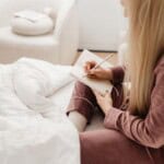A person in mauve pajamas sits on a bed, journaling for beginners in a notebook with a pen. White bedding and a soft chair are visible, creating a cozy, peaceful atmosphere.