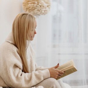 A woman with long blonde hair, wearing a cozy cream-colored fleece outfit, sits indoors reading a book, finding comfort much like journaling when feeling overwhelmed. The background features soft lighting and blurred neutral decor.