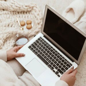 A person sitting on a cozy blanket with a laptop on their lap, holding it with both hands—making journaling fun. Nearby is a plate of cookies and a cup of coffee or tea resting on the floor.