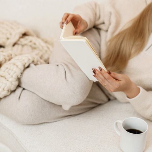A person in cozy beige loungewear sits on a white couch with a chunky knit blanket, reading their first journal entry. A mug of coffee rests nearby on the couch. The scene feels calm and relaxed.