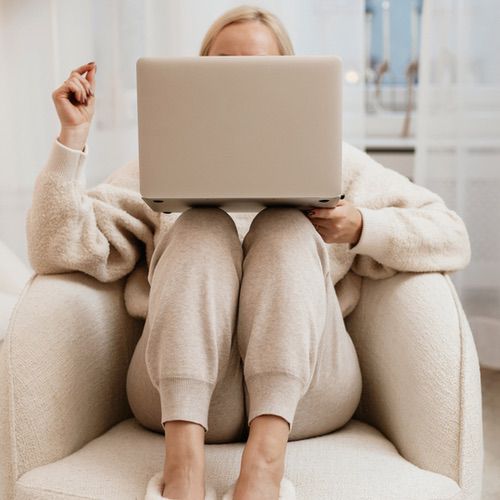 A person in cozy, light-colored loungewear sits in a plush armchair with legs folded up, holding a laptop on their knees—perhaps typing their first journal entry, their face mostly hidden behind the screen.