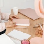 A person with pink nail polish holds a closed pink notebook at a tidy wooden desk, featuring a pink pen, open notebook with guided journal prompts for mental health, pink laptop, glasses, coffee cup, candle, and desk organizer.