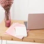 A light wooden desk with a pink laptop, white notebook, pink folders, a pen, a pink candle, and a vase of small pink flowers—perfect inspiration if you’re learning how to make a guided journal in a soft, feminine workspace.