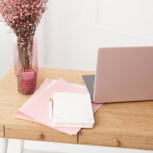 A light wooden desk with a pink laptop, white notebook, pink folders, a pen, a pink candle, and a vase of small pink flowers—perfect inspiration if you’re learning how to make a guided journal in a soft, feminine workspace.