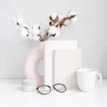 A minimalist desk scene features a white mug, round glasses, two closed books with morning guided journal prompts, a small white container, and a modern pink vase holding cotton stems, all set against a light background.