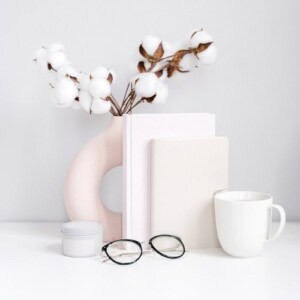 A minimalist desk scene features a white mug, round glasses, two closed books with morning guided journal prompts, a small white container, and a modern pink vase holding cotton stems, all set against a light background.