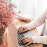 A person wearing a light pink sweater types on a laptop at a wooden desk, where a self care guided journal, a vase of pink flowers, and notebooks add to the soft, pastel aesthetic.