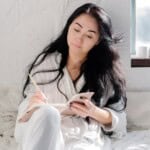 A woman with long black hair, wearing a white robe, sits on a bed by a sunlit window, writing thoughtfully in her stress relief journal with a pencil.