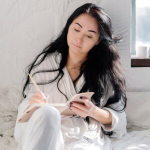 A woman with long black hair, wearing a white robe, sits on a bed by a sunlit window, writing thoughtfully in her stress relief journal with a pencil.