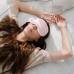A woman with long brown hair is lying on a bed wearing a light pink sleep mask and a beige shirt, one arm resting above her head on the pillow, finding peace after an evening of journaling for mental health.