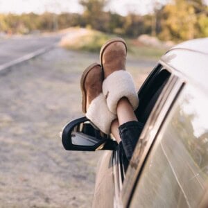 A confident woman wearing tan shearling boots rests her legs out of a car window, framed by a scenic outdoor backdrop of trees and an empty road.