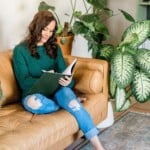 A woman with long brown hair, wearing a green sweater and ripped jeans, sits on a tan couch reading a book filled with stress management journaling prompts. She is surrounded by large green houseplants in a cozy, well-lit living room.