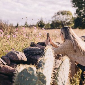 A woman in a beige sweater kneels by a stone wall, taking a photo of wildflowers with her phone in a sunny, rural field—showing how nature can inspire rather than destroy your confidence. Cacti and greenery fill the vibrant foreground.