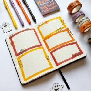 An open bullet journal with halloween-themed decorations, featuring orange and yellow bordered boxes, halloween journal supplies like washi tape, pens, and ghost stickers arranged around the notebook.
