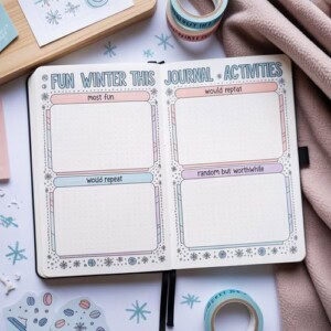 An open journal displays a winter-themed activities spread with four labeled boxes for winter journal activities: most fun, would repeat, would repeat, and random but worthwhile. Washi tape rolls, paper, and a pink blanket surround the journal.