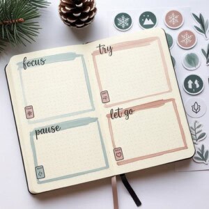 An open bullet journal displays four labeled sections—focus, pause, try, and let go—surrounded by pine branches, a pine cone, and round winter-themed stickers, offering cozy winter journal ideas for inspiration.