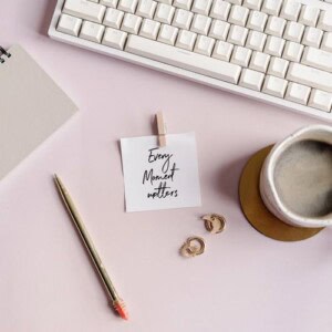 A white keyboard, a notebook for free writing, a pen, a pair of earrings, a coffee cup, and a note with “every moment matters” handwritten on it are arranged on a light pink desk.