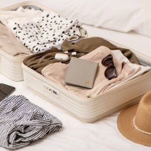 Open cream-colored suitcase with neatly folded clothes, a pair of sunglasses, a beige passport holder, and wireless earbuds inside. Nearby are a striped shirt, straw hat, tablet, and tips on how to travel journal resting on a white bed.