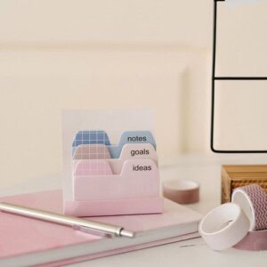 A set of pastel-colored file tabs labeled notes, goals, and ideas sits on a pink notebook—perfect for reflection journaling—alongside a silver pen and rolls of decorative tape on a desk.
