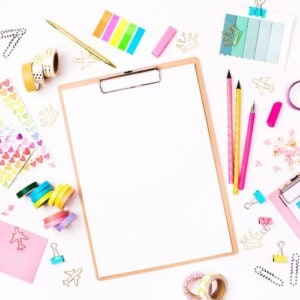 A blank clipboard is centered on a white desk surrounded by colorful stationery items, perfect for color coding your planner or journal with sticky notes, washi tape, pencils, pens, paper clips, stickers, and paint swatches.