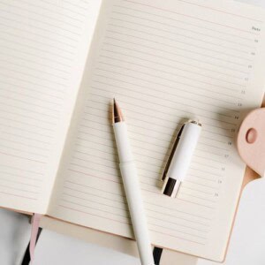 An open lined notebook with dated pages lies on a desk, perfect as a guide to dream journaling. A white and gold pen rests on the notebook, surrounded by soft pink décor and other office supplies in the background.