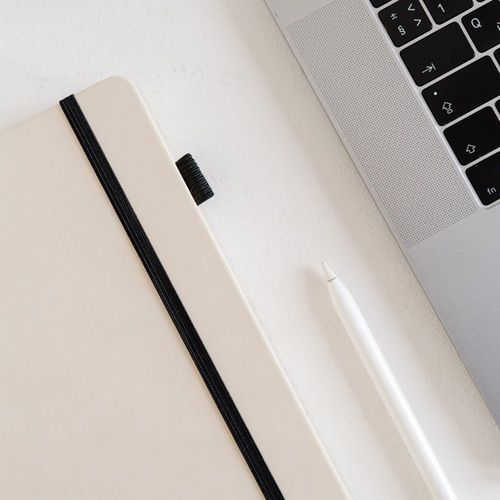 A closed beige notebook with a black elastic band, a white stylus, and part of a laptop keyboard rest on a white surface—perfect tools for those learning how to gratitude journal.