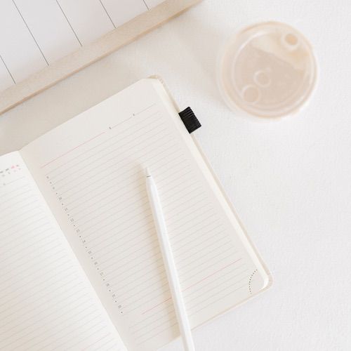 A lined notebook open to a blank page with a white pen resting on it—perfect for starting your how to gratitude journal—sits beside a plastic cup of light-colored drink, all arranged on a clean white surface.