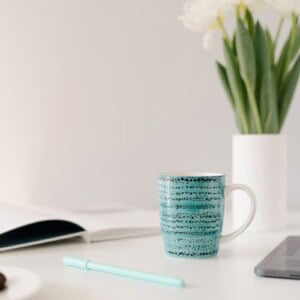 A turquoise mug sits on a white desk next to an open notebook—perfect for keeping a commonplace book—a teal pen, a laptop edge, and a vase of white flowers in the background.