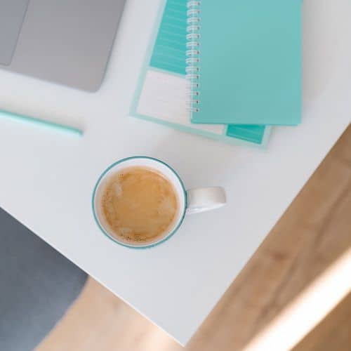 A cup of coffee sits on a white desk beside a turquoise notebook—perfect for keeping a commonplace book—a matching pen, and part of a silver laptop. The scene has a bright, minimalist feel with light wooden flooring visible.
