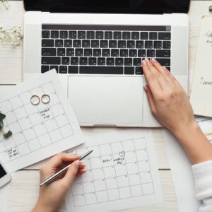 A person uses a laptop while holding a pen and marking a calendar, perhaps exploring types of planners for journaling. Two rings rest on another calendar amid papers, a phone, and a notebook on the white desk.