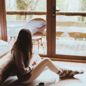 A woman with long hair in a hoodie and patterned socks sits on the floor by a large glass door, seeking calm after an anxiety attack as she looks outside at a wooden deck and trees. A cup and an open notebook rest beside her.