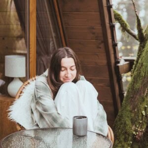 A woman sits on a cozy chair outside a wooden cabin, hugging her knees and smiling with her eyes closed. Wrapped in a blanket, she enjoys journaling for anxiety, with a mug on the table and mossy trees in the background.