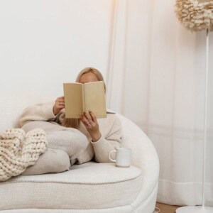 A person in cozy, neutral loungewear sits on a white sofa, reading a book to soothe your anxiety. A chunky knit blanket covers their legs, and a white mug rests nearby. The calm scene features soft lighting and minimal, warm decor.