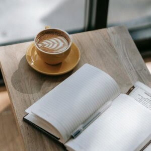 A cup of latte with latte art sits on a yellow saucer next to an open blank notebook and pen, ready for jotting down journal prompts for emotional healing, on a wooden table by a window.