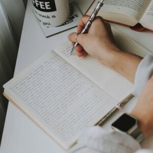 A person writes in a lined notebook with a pen, wearing a smartwatch. An open book and coffee mug sit on the white desk beside them as they reflect on journal prompts for physical healing.