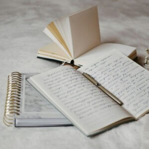 Three notebooks are arranged on a surface: one open with handwritten notes in cursive, one spiral-bound notebook, and one book with blank pages fanned open—perfect for mood and emotion journal prompts. A pen rests on the open notebook.