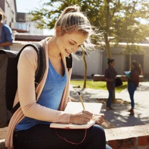 A young woman with a rucksack sits outdoors, smiling as she writes in her notebook, journaling for self-esteem and confidence. Two other people stand and talk in the background near trees and a building.