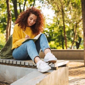 A young woman with curly red hair, wearing a yellow jumper and jeans, sits outdoors on a bench, smiling as she writes self-esteem and confidence journal prompts in her notebook. Green trees and sunlight create a peaceful park setting.